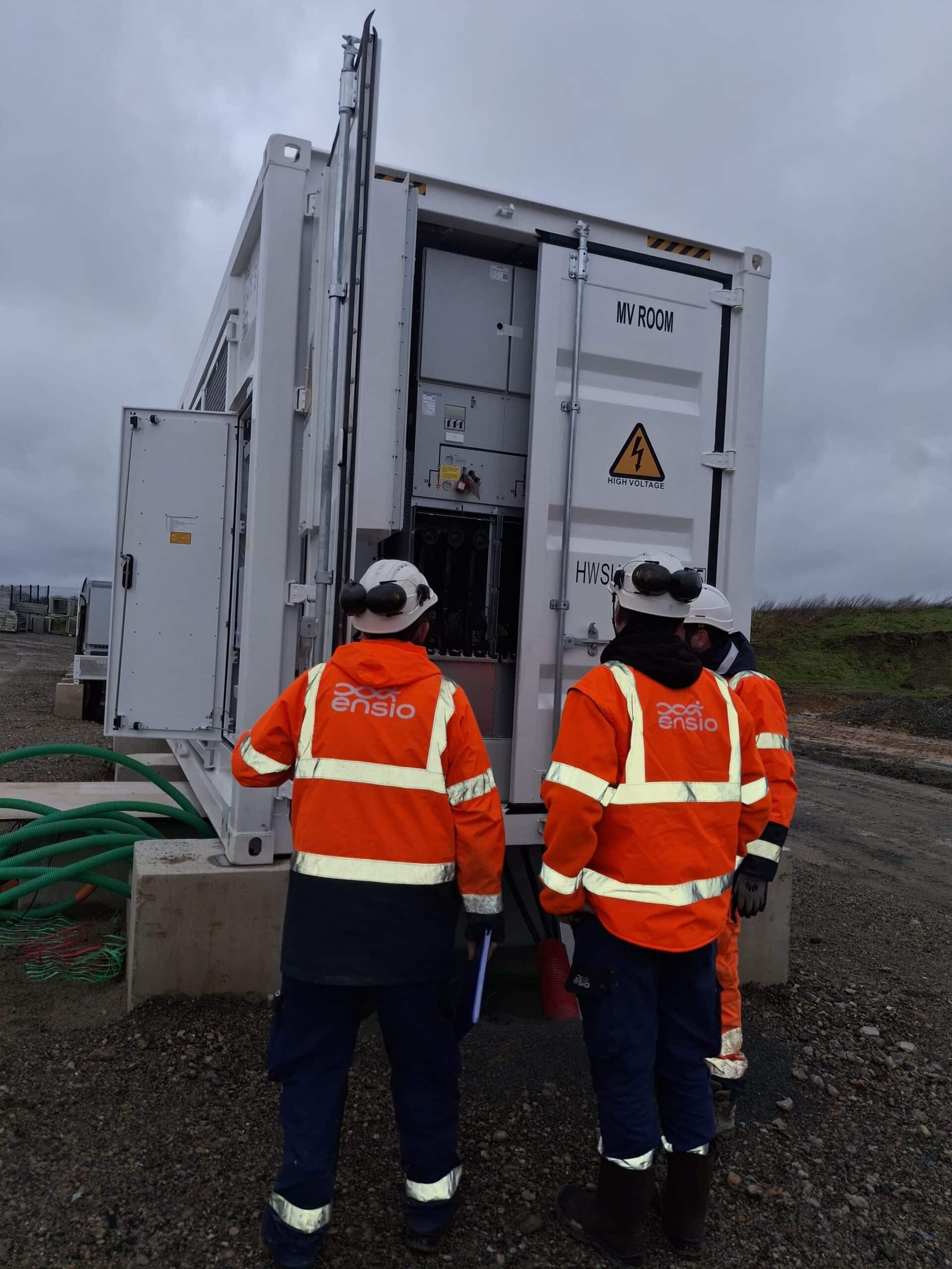 Deux techniciens ENSIO de dos en veste orange inspectant l'intérieur d'un container de stockage d'énergie moyenne tension sur un chantier BESS