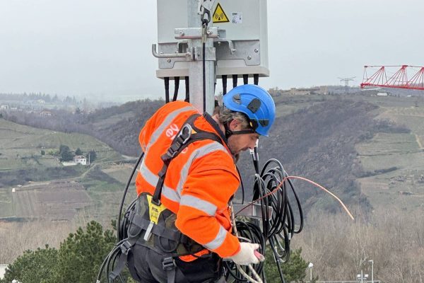 Technicien en veste orange et casque bleu équipé d'un harnais intervenant en hauteur sur un mât télécom avec câbles, sur fond de paysage vallonné